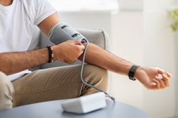 A man sitting on a couch at his home places a blood pressure monitoring cuff on his upper arm